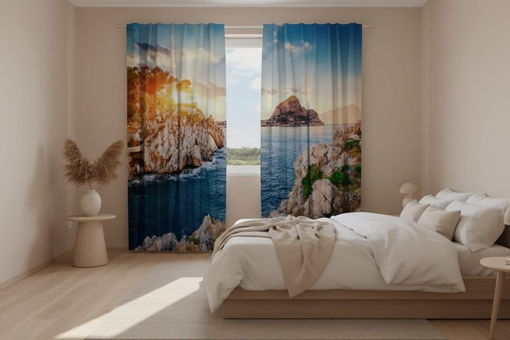 Gaišas guļamistabas interjers ar baltām sienām un koka grīdu. Uz loga uzstādīti fotoaizkari, uz kuriem fotoreālistiski attēlota klinšaina Vidusjūras ainava (Sicīlijas piekraste). Kreisajā aizkaru panelī redzamas stāvas, gaišas klintis ar zaļiem kokiem virsotnē, aiz kuriem spīd spilgta, zeltīta saules gaisma. Labajā panelī redzams mierīgs, tumši zils jūras ūdens, akmeņains krasts priekšplānā un liela klinšu sala fonā.