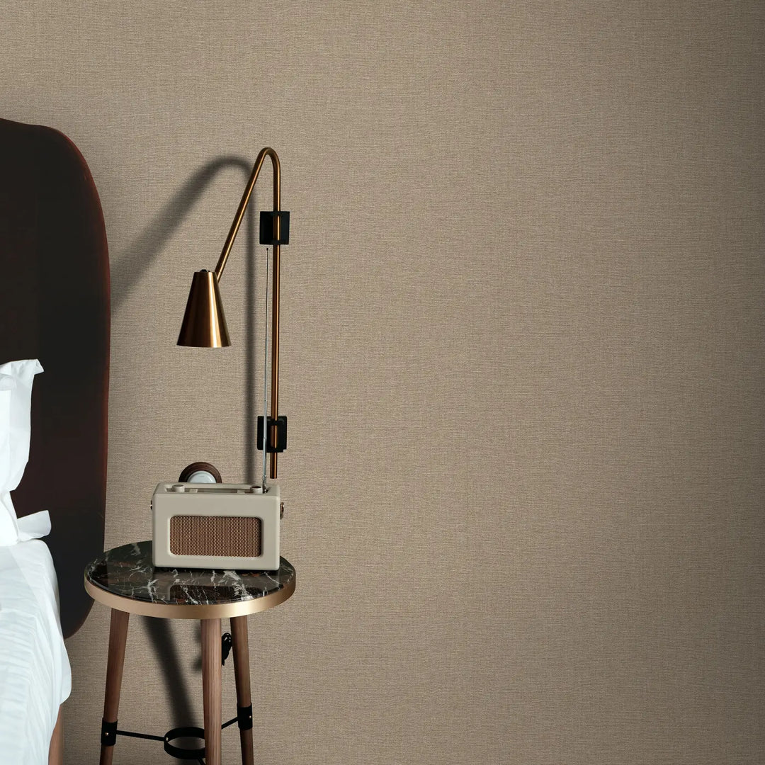 Silti bēšas tekstūras tapetes ar lina auduma efektu guļamistabas interjerā. Priekšplānā elegants naktsskapītis ar tumša marmora virsmu, uz kura atrodas retro stila radio un moderna misiņa sienas lampa.