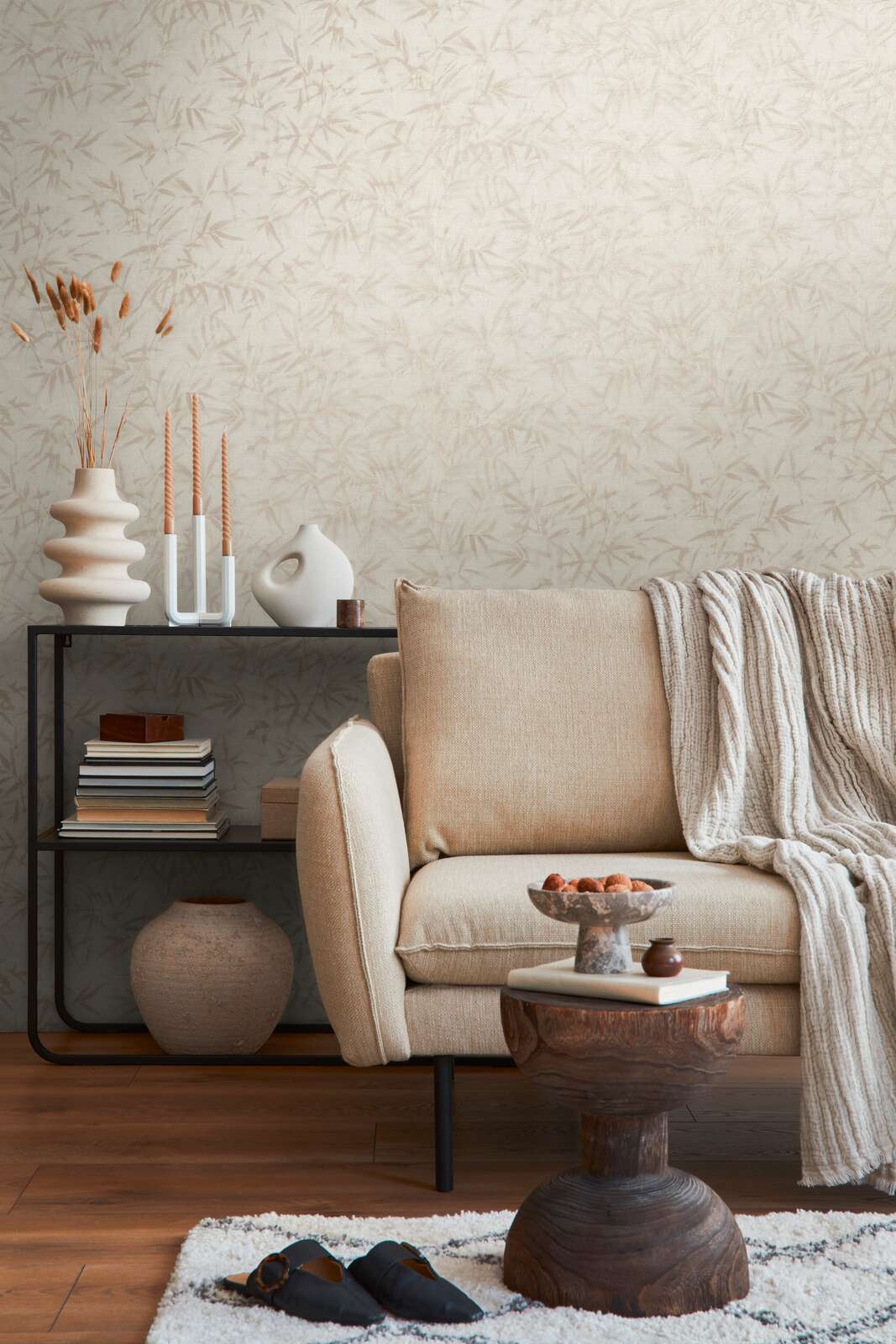 Light cream bamboo leaf pattern wallpaper in an interior with a light sofa, black shelving unit, and brown wood plank flooring with a light rug