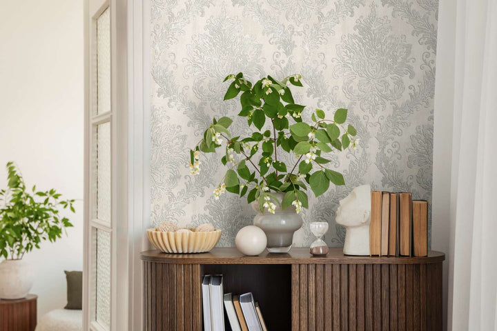 Light gray damask pattern wallpaper in an interior behind a wooden cabinet with a plant in a vase and decor