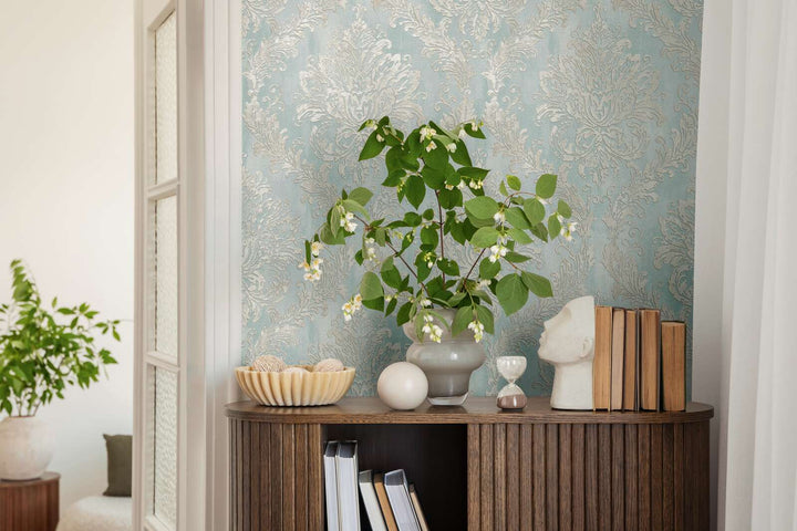 Light blue damask pattern wallpaper in an interior behind a wooden cabinet with a plant in a vase and decor