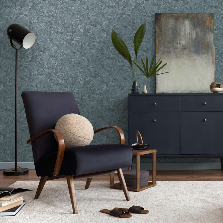 Teal bamboo leaf pattern wallpaper in an interior with a black lounge chair, dark dresser, and natural wood plank flooring with a light rug