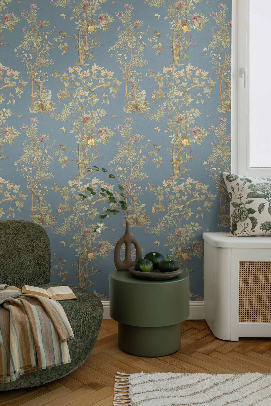 Dark blue illustrated nature motif wallpaper in an interior with a green lounge chair, round side table, and natural wood parquet flooring with a rug