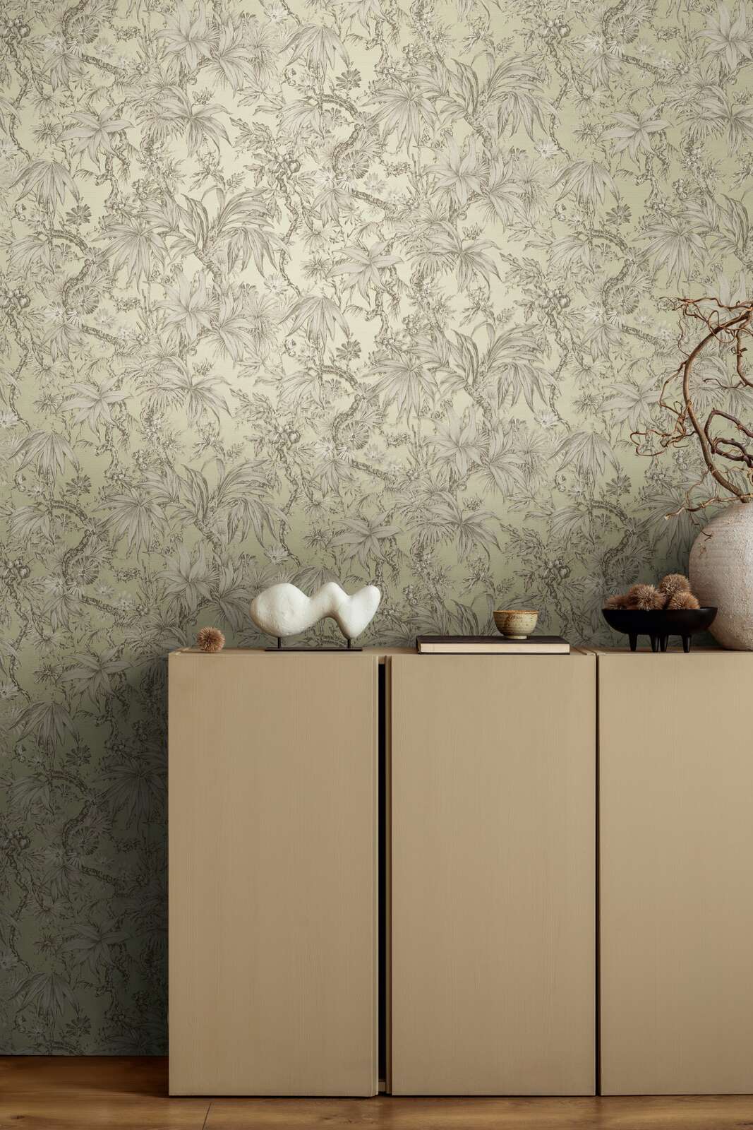 Light greenish botanical pattern wallpaper in an interior behind a beige dresser with decor and brown wood plank flooring